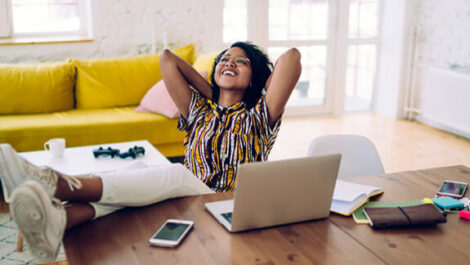 A woman is sitting at her table in front of her laptop, relaxing with her hands behind her head.