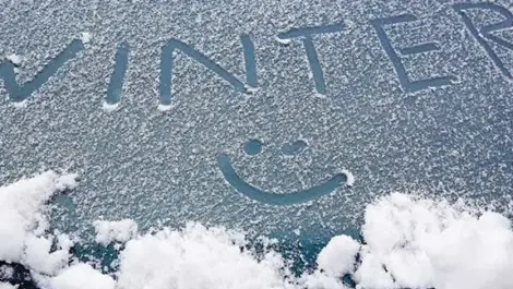 A car's window has the word "Winter" spelled out in the snow that has accumulated on top of it. There is also a smiley face drawn below the word.