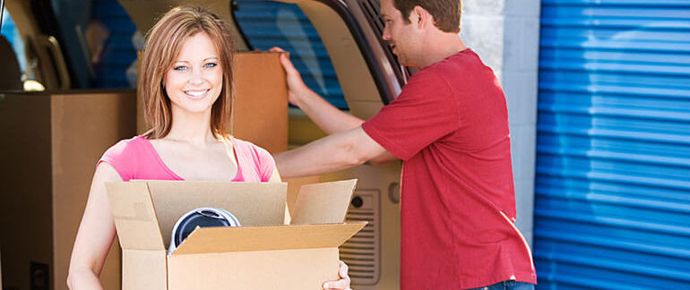 Woman carrying moving box while man loads boxes into vehicle.