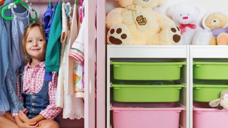 Child smiling in clothing closet surrounded by clothes and storage.