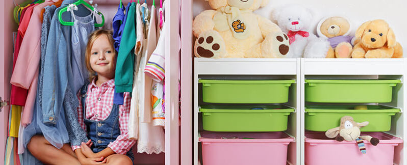 Child smiling in clothing closet surrounded by clothes and storage.