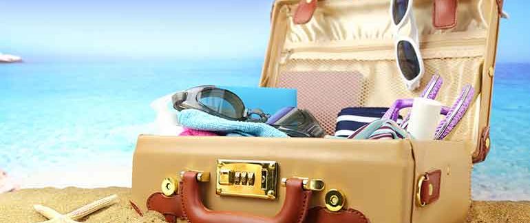 Open suitcase on a beach that houses goggles, towels, and sunglasses.