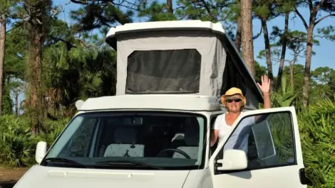 A woman waves to the camera, about to enter her camper van