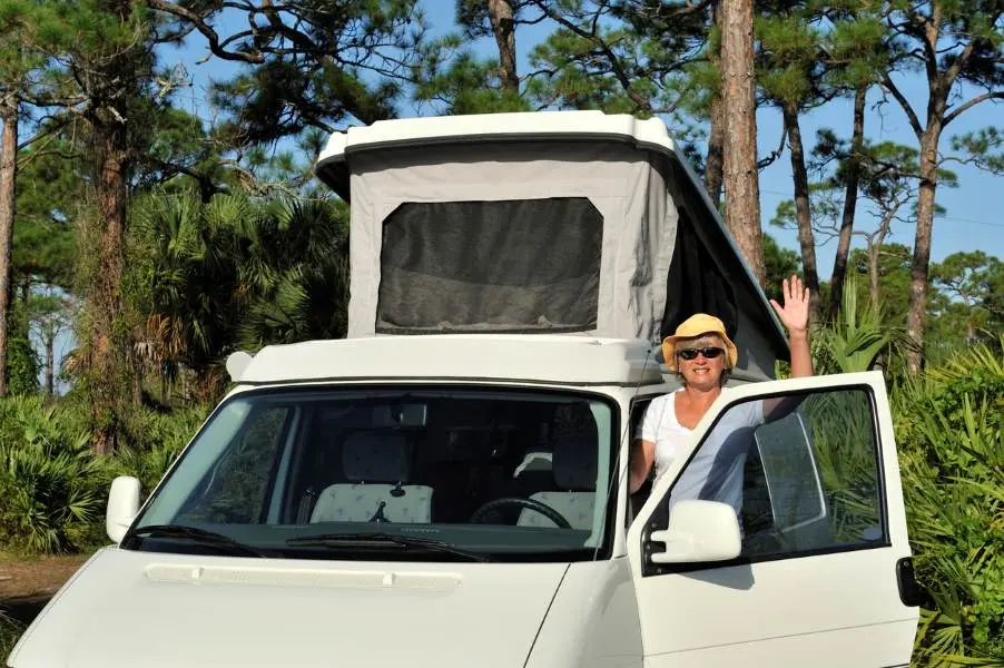 A woman waves to the camera, about to enter her camper van