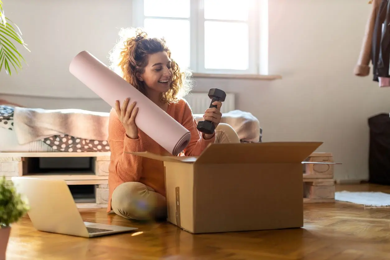 A woman with curly hair wearing workout clothes smiles and sits on the floor opening package with a yoga mat and dumbbell inside