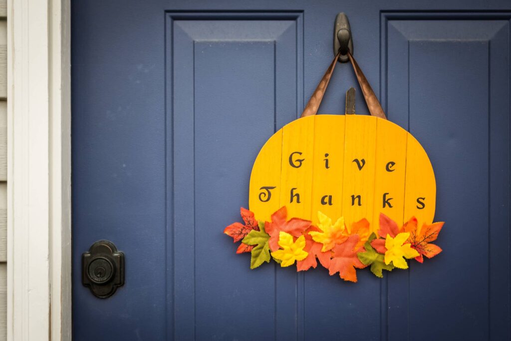 An orange wooden sign shaped like a pumpkin hangs on a door, reading "Give Thanks." 