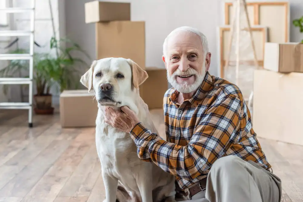 Elderly man sitting on the floor next to his dog in front of several stacks of boxes.