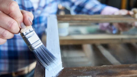 A person wearing plaid uses a paintbrush to give a wooden frame a coat of white paint.