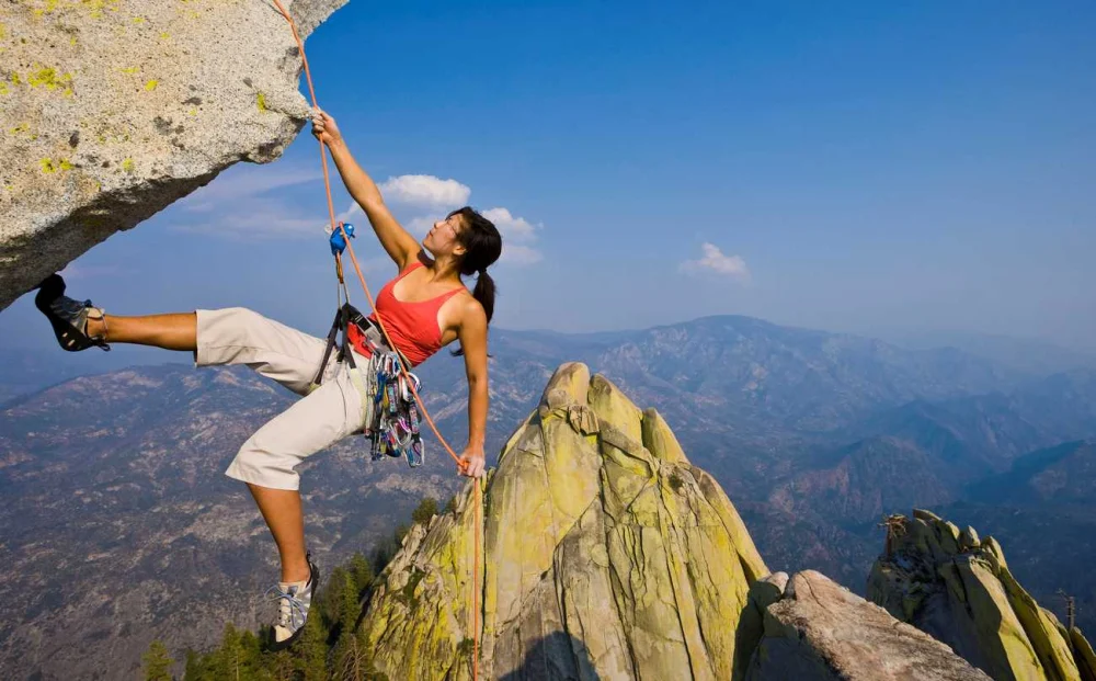 A female rock climber wearing a red tank top hangs from a high cliff