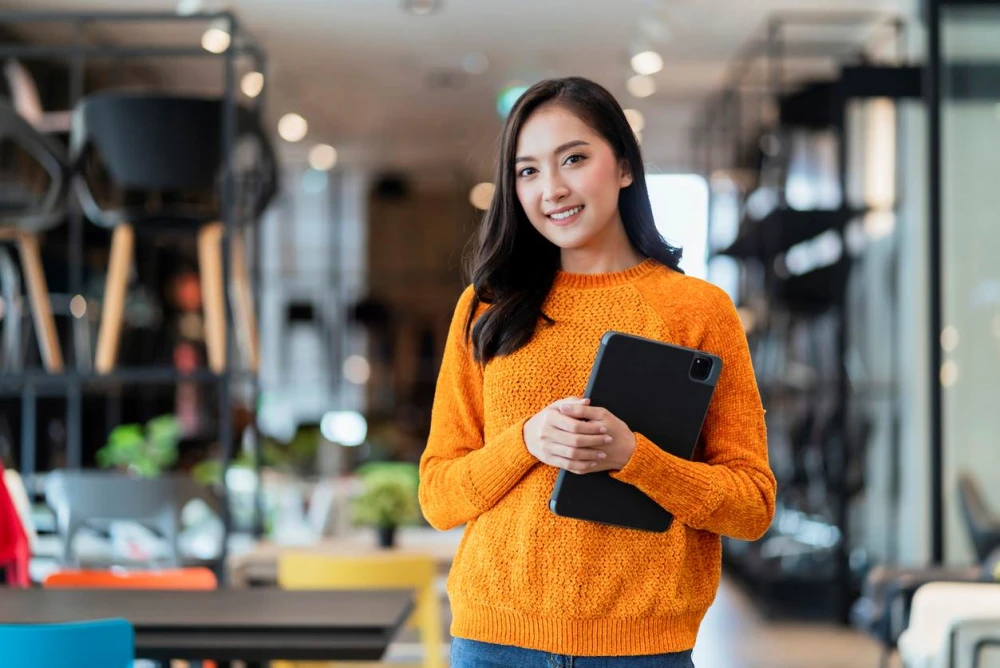 A smiling businesswoman stands in a workspace holding a tablet