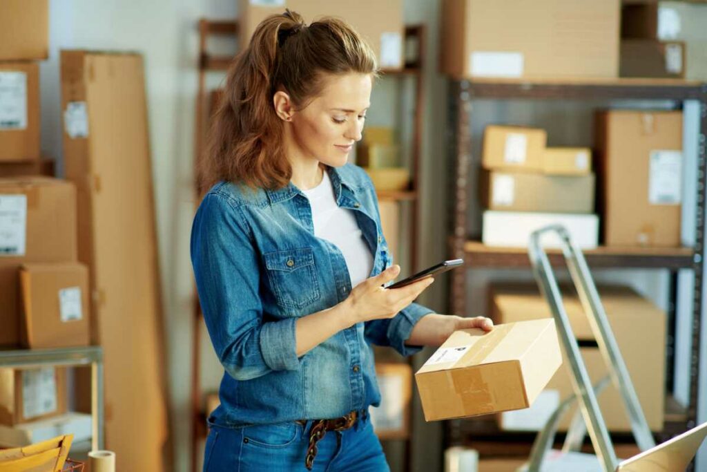 A small business owner uses her phone in a storage unit surrounded by boxes.