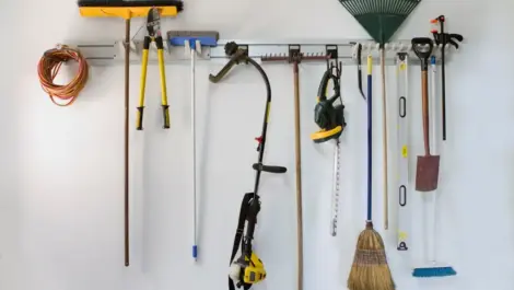 Common gardening tools, including a rake, shovel, chainsaw, and weed whacker, hang on a rack.