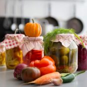 A closeup view of canned goods in glass jars with a pile of vegetables in front.