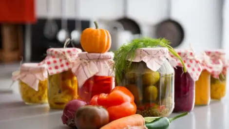 A closeup view of canned goods in glass jars with a pile of vegetables in front.