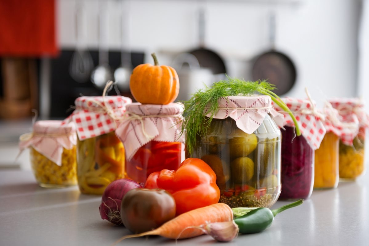 A closeup view of canned goods in glass jars with a pile of vegetables in front.