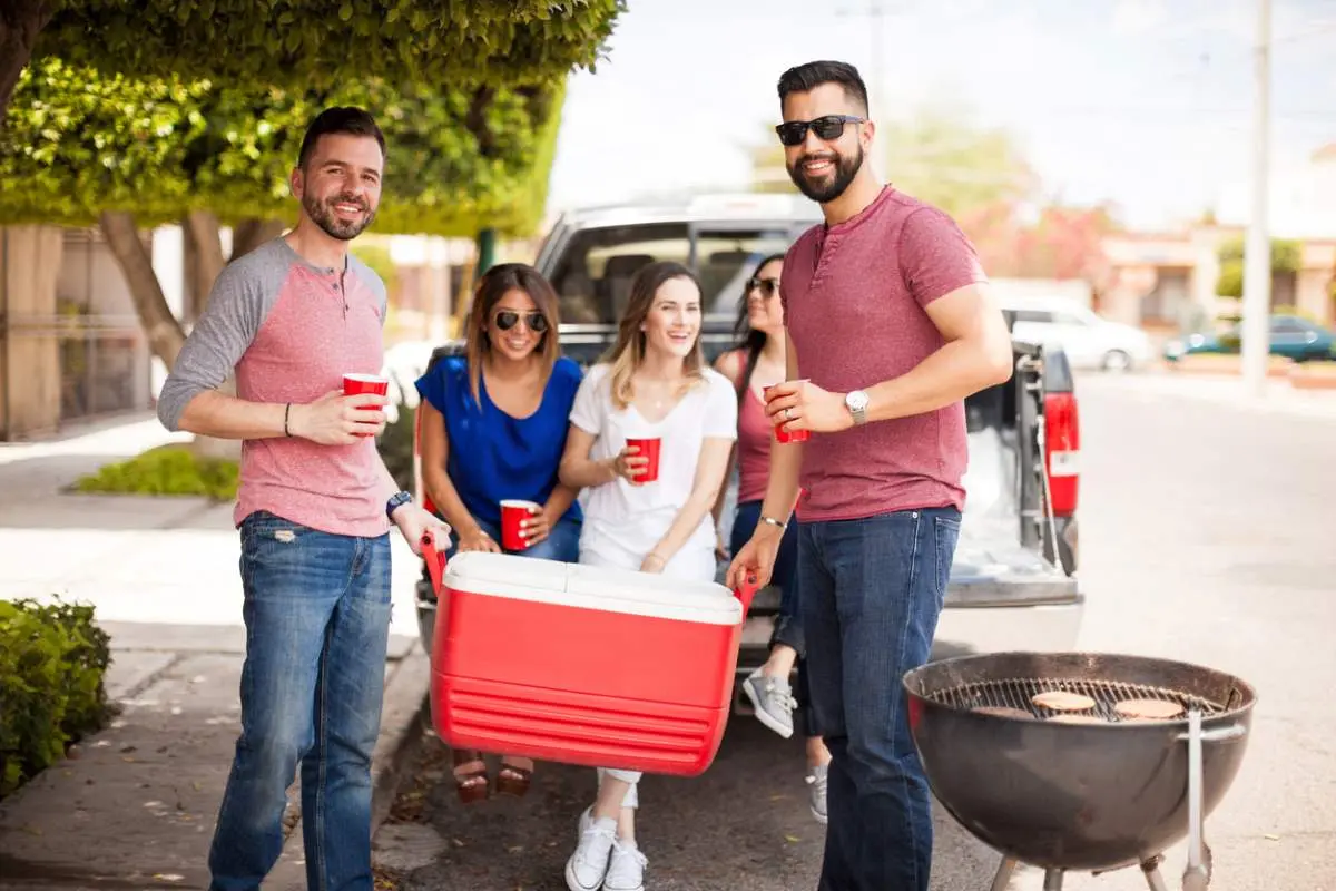 Two men hold a red cooler next to a grill as others smile in the background. 