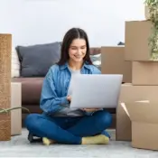 A college-aged girl sits amid boxes and belongings in her living space while using a laptop.