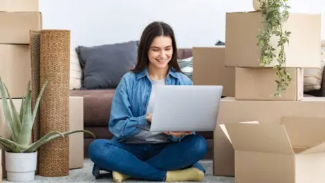 A college-aged girl sits amid boxes and belongings in her living space while using a laptop.