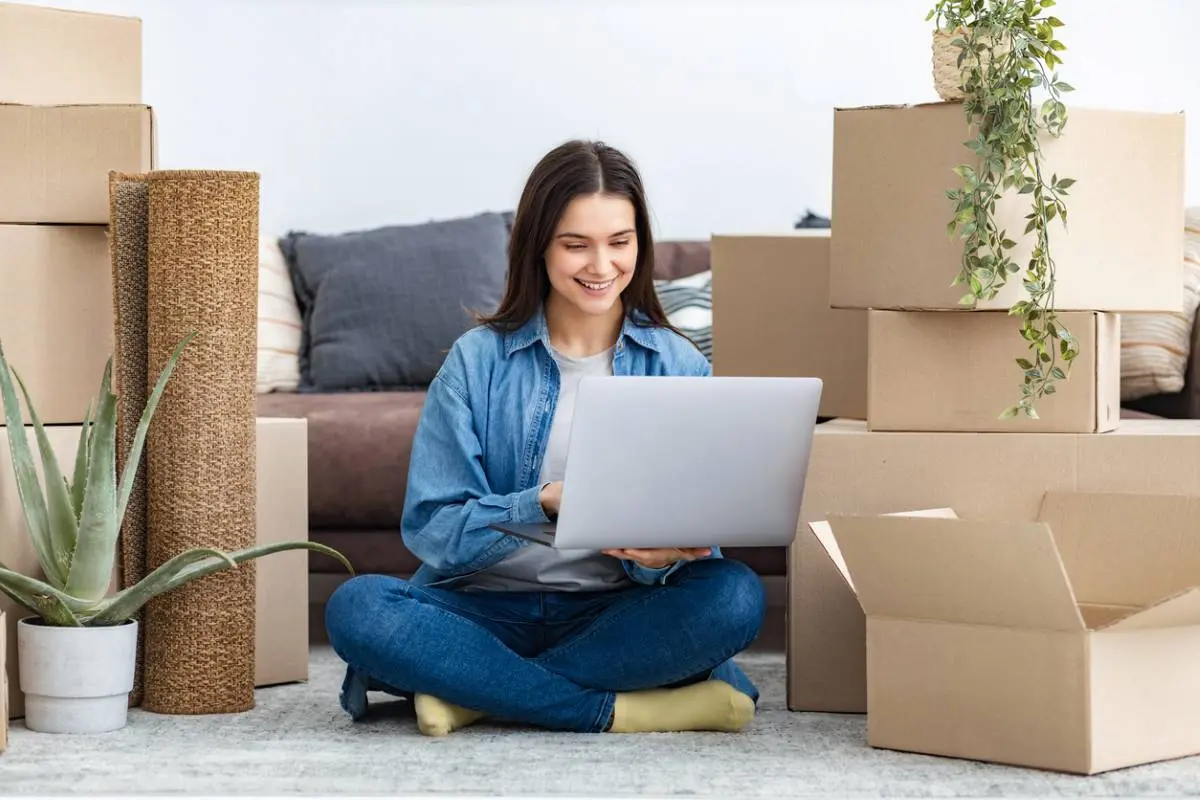 A college-aged girl sits amid boxes and belongings in her living space while using a laptop.