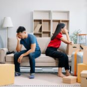 A divorcing couple sit on opposite sides of the couch amid moving boxes.