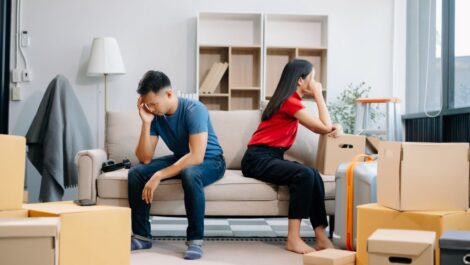 A divorcing couple sit on opposite sides of the couch amid moving boxes.