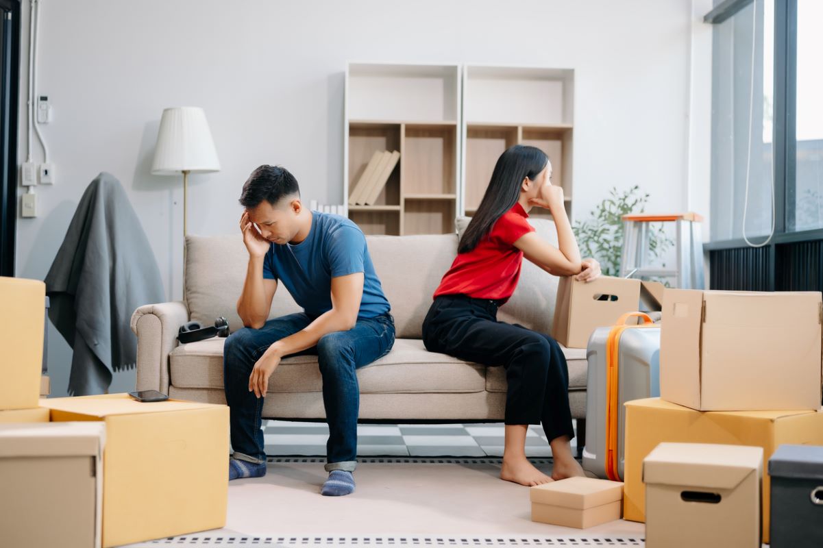 A divorcing couple sit on opposite sides of the couch amid moving boxes.