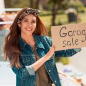 A smiling young woman in a jean jacket holds a garage sale sign.