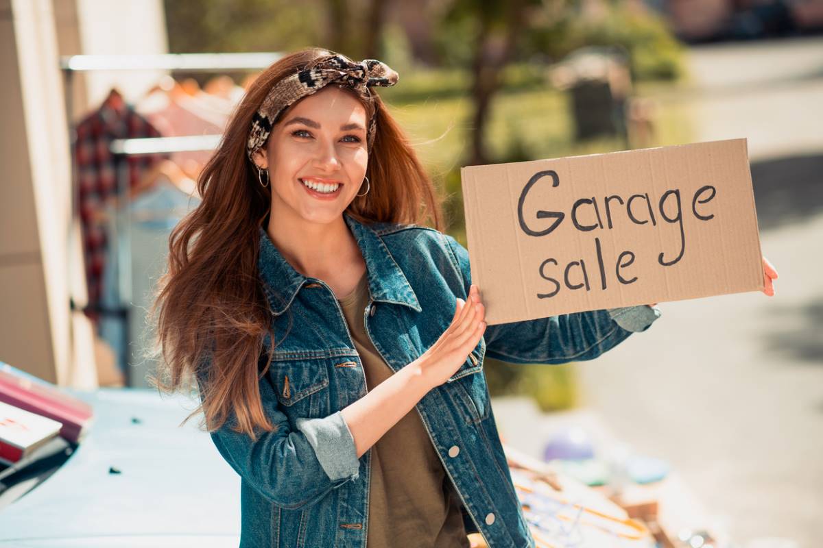 A smiling young woman in a jean jacket holds a garage sale sign.