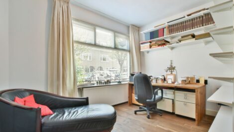 A spare bedroom used as an office has a desk, office chair, shelves, and lounge chair.