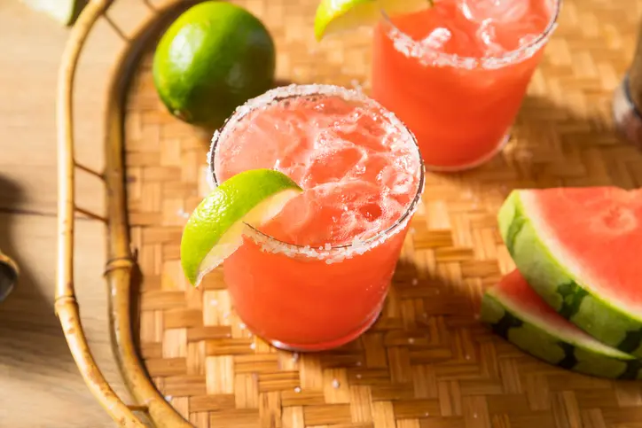 Two watermelon margaritas with a lime garnish sit on a woven tray with a lime and watermelon slice beside. 