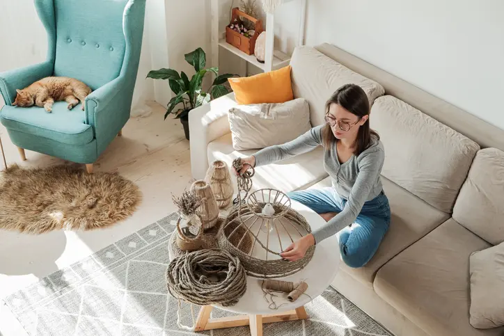 A young woman makes a DIY lamp while sitting on a beige couch with a teal armchair with a sleeping orange cat beside. 
