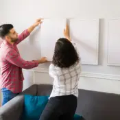 A young couple hangs a plain white canvas on the wall next to two other plain canvases.
