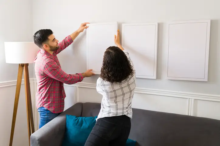 A young couple hangs a plain white canvas on the wall next to two other plain canvases.
