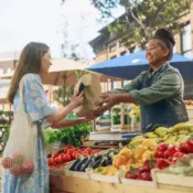 A smiling vendor hands a lady in a blue dress a paper bag of produce at a farmers market